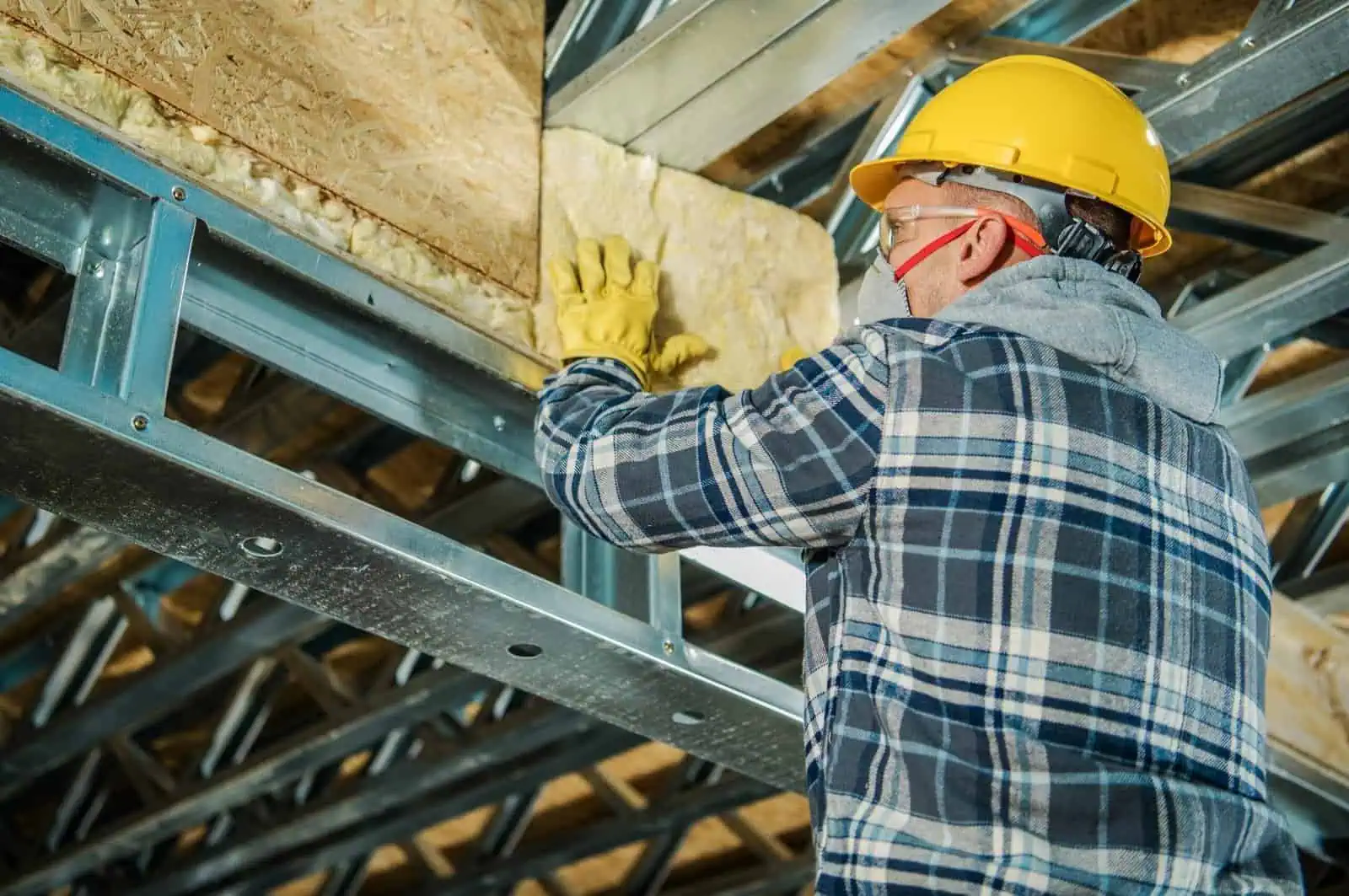 Worker installing insulation in a building ceiling with safety gear.