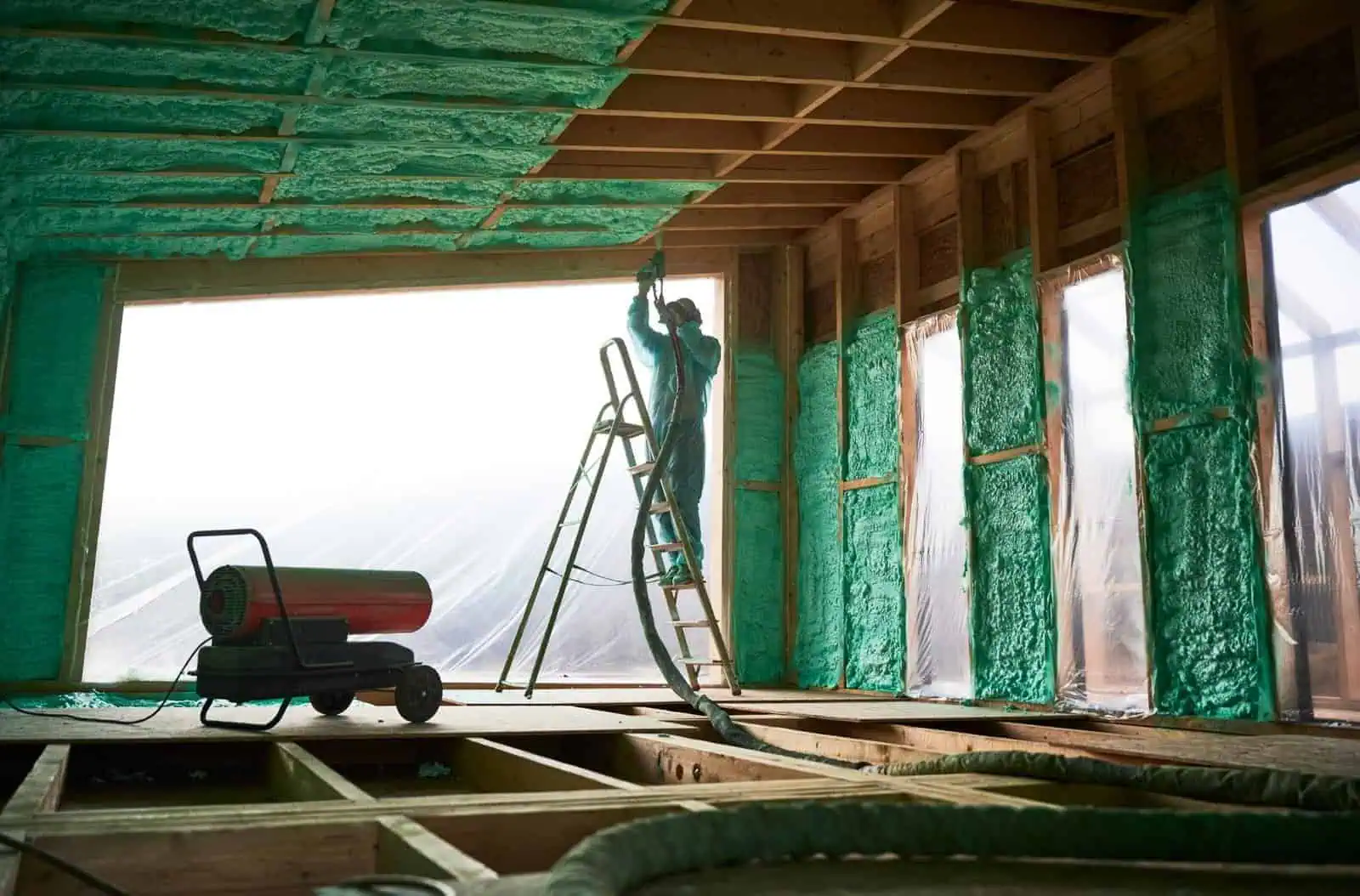 Worker applying green spray foam insulation to wall in construction site.
